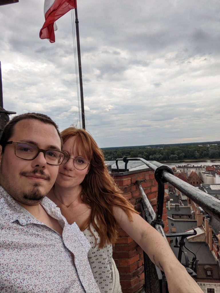 Selfie d'Alicia et Florent pendant leur roadtrip en Pologne en haut de la tour de l'hôtel de ville de Toruń avec le drapeau polonais en arrière-plan © Le Voyage de FloLili - Blog de Voyage